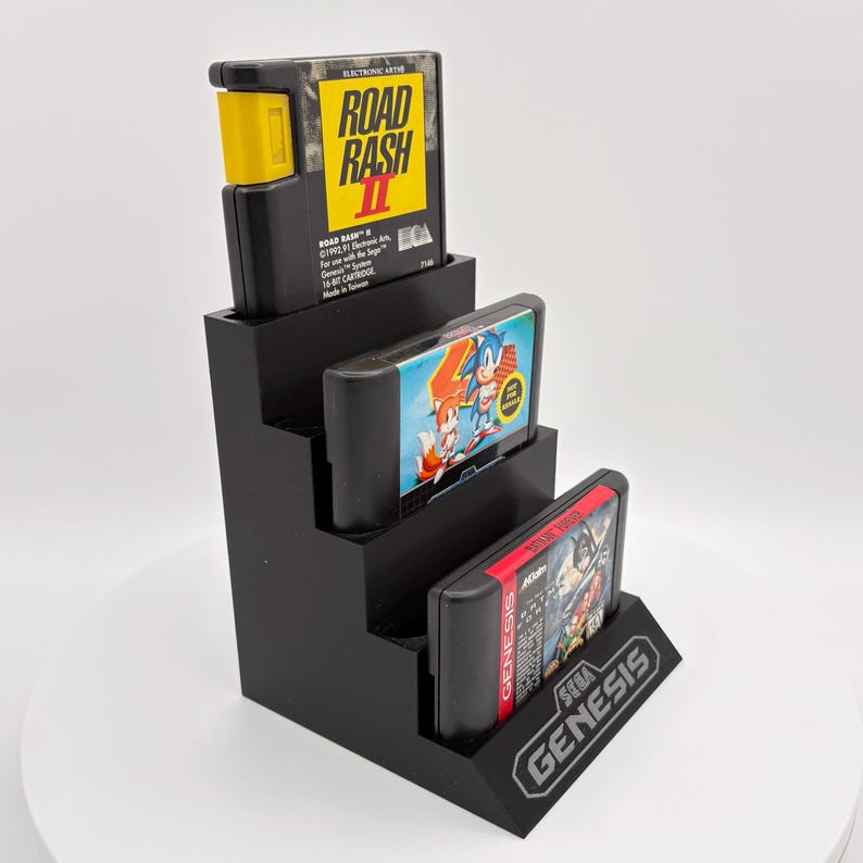 K&ouml;nnte beinhalten: Ein schwarzer, stufenf&ouml;rmiger Displayst&auml;nder mit drei Retro-Videospielkassetten. Die oberste Kassette ist mit "Road Rash II" beschriftet, die mittlere zeigt Sonic the Hedgehog und die untere ist mit "Sega Genesis" beschriftet.