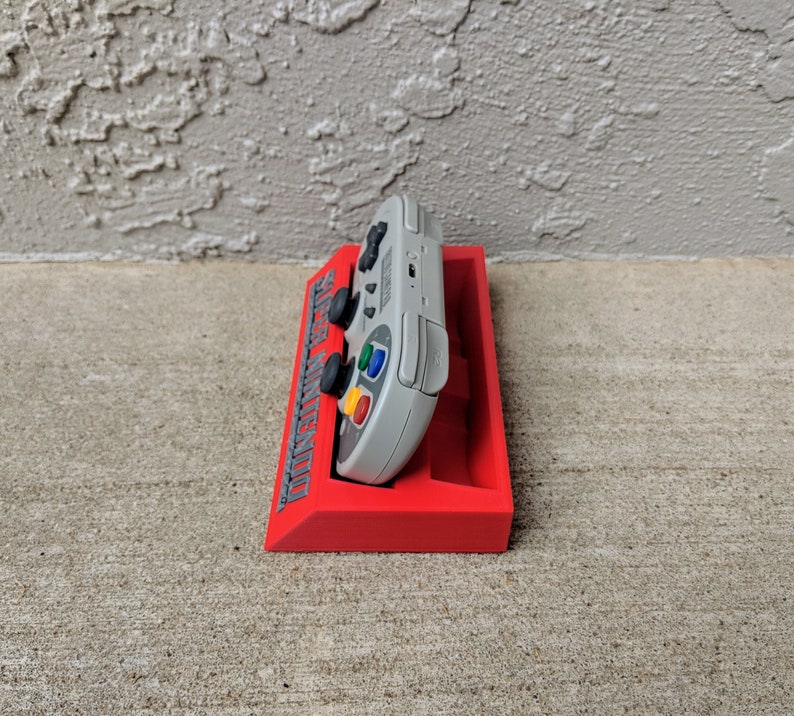May include: A red plastic stand with the words "Super Nintendo" printed on the side. A gray Super Nintendo controller is resting in the stand.