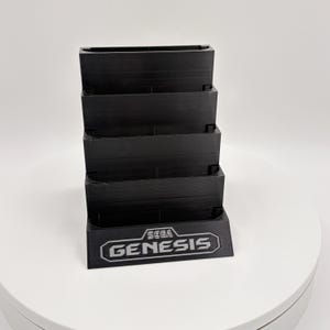 K&ouml;nnte beinhalten: Ein schwarzer Spielkassettenhalter mit mehreren Ebenen, mit dem Sega Genesis-Logo in Wei&szlig;. Der Halter dient zur Aufbewahrung von Spielkassetten. Der Halter besteht aus einem soliden, mattschwarzen Material.