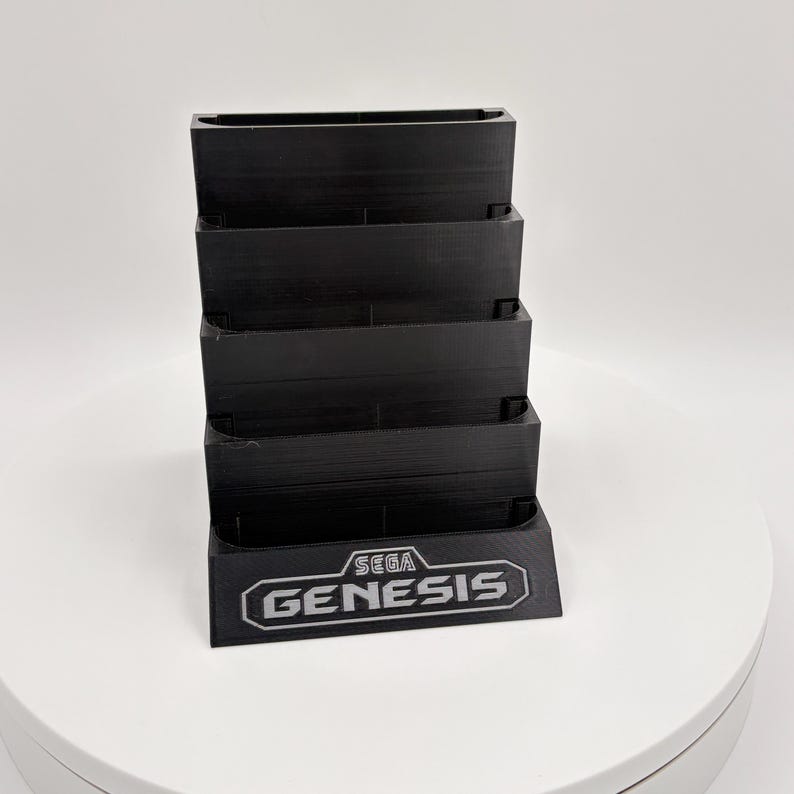 K&ouml;nnte beinhalten: Ein schwarzer Spielkassettenhalter mit mehreren Ebenen, mit dem Sega Genesis-Logo in Wei&szlig;. Der Halter dient zur Aufbewahrung von Spielkassetten. Der Halter besteht aus einem soliden, mattschwarzen Material.