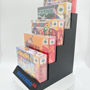 May include: A black, stepped display stand holding five Nintendo 64 game boxes. The games include "The Legend of Zelda", "Star Fox 64", "Mario Party 3", and "Super Smash Bros". The stand has the text "NINTENDO 64" on the front.