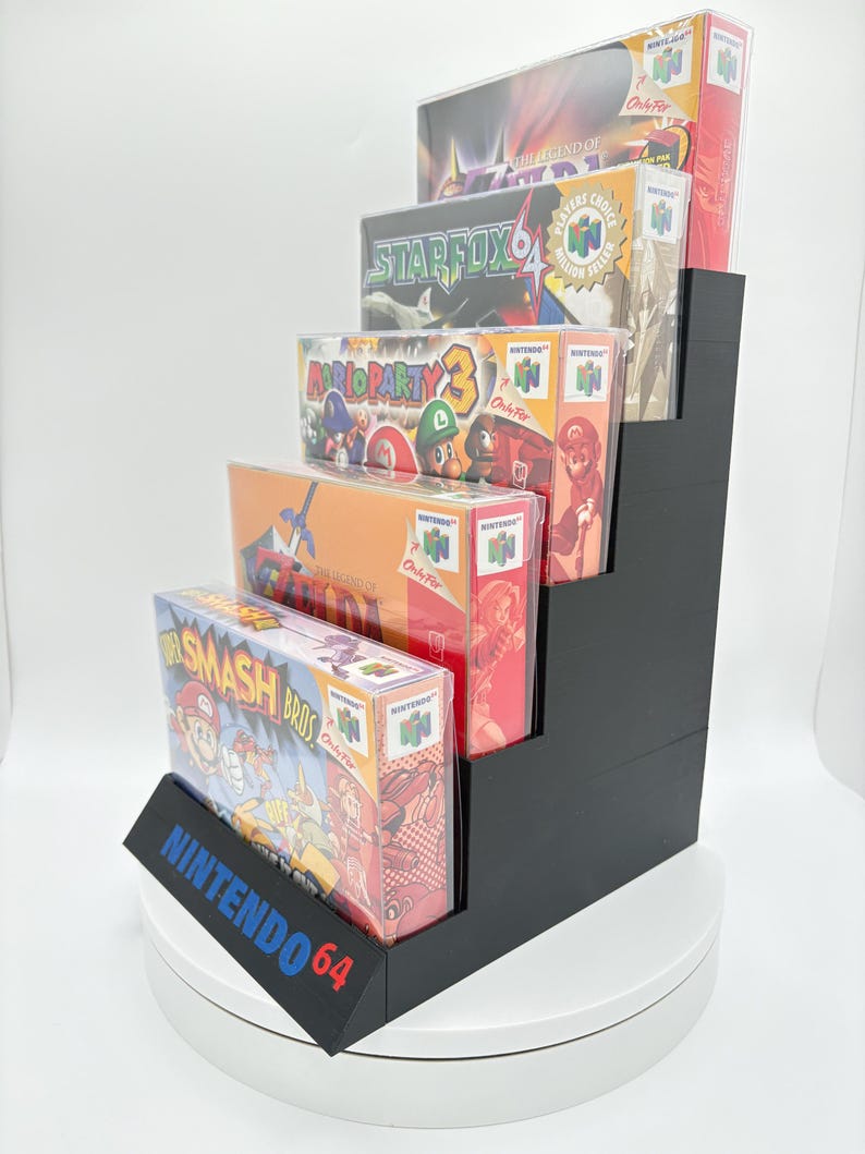 May include: A black, stepped display stand holding five Nintendo 64 game boxes. The games include "The Legend of Zelda", "Star Fox 64", "Mario Party 3", and "Super Smash Bros". The stand has the text "NINTENDO 64" on the front.