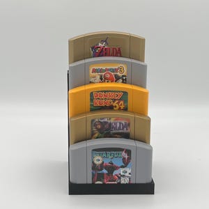 May include: A black stand with four slots holds five Nintendo 64 game cartridges. The cartridges are from top to bottom: The Legend of Zelda: Ocarina of Time, Mario Party 3, Donkey Kong 64, The Legend of Zelda: Majora's Mask, and Star Fox 64.