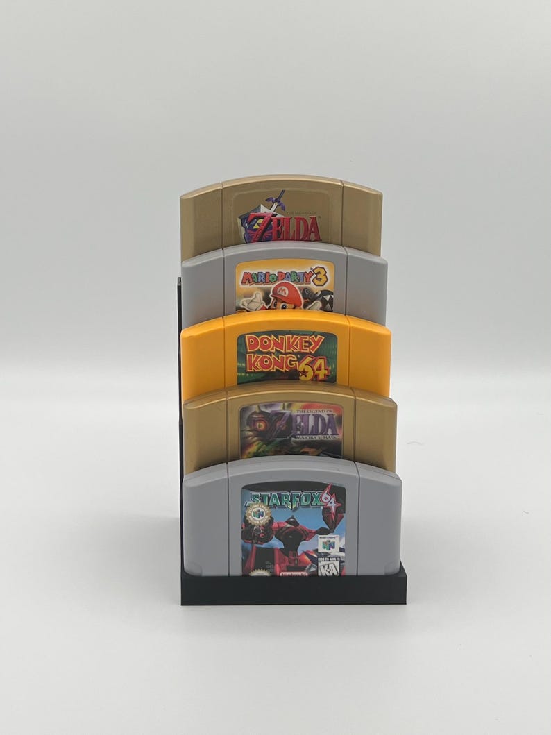 May include: A black stand with four slots holds five Nintendo 64 game cartridges. The cartridges are from top to bottom: The Legend of Zelda: Ocarina of Time, Mario Party 3, Donkey Kong 64, The Legend of Zelda: Majora's Mask, and Star Fox 64.