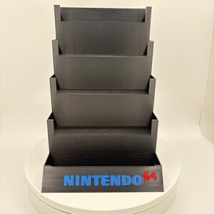 May include: Black tiered storage unit for video game cartridges. The unit has five shelves and features the blue text "NINTENDO" with "64" in red. The unit is designed to hold and display video game cartridges.