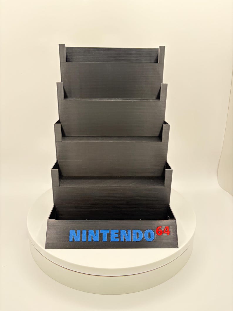 May include: Black tiered storage unit for video game cartridges. The unit has five shelves and features the blue text "NINTENDO" with "64" in red. The unit is designed to hold and display video game cartridges.