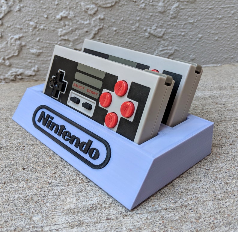 May include: A purple plastic stand with the word "Nintendo" printed on the front. Two gray Nintendo Entertainment System controllers are sitting in the stand.