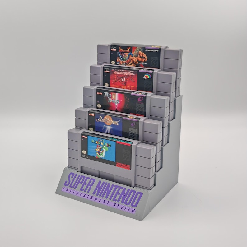 Puede incluir: Un soporte de pl&aacute;stico gris con una etiqueta morada que dice "SUPER NINTENDO ENTERTAINMENT SYSTEM". El soporte contiene cinco cartuchos de juego de Super Nintendo, incluyendo "Mortal Kombat", "The Secret of Mana", "Chrono Trigger", "Super Mario World" y "Street Fighter II: The World Warrior".