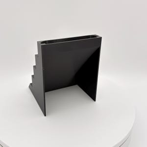 K&ouml;nnte beinhalten: Ein schwarzer, geometrischer Pr&auml;sentationsst&auml;nder mit einem Stufendesign auf einer Seite und einer flachen Oberfl&auml;che auf der anderen Seite. Der St&auml;nder hat eine rechteckige Oberseite und besteht aus einem soliden, undurchsichtigen Material. Das Design ist modern und minimalistisch.