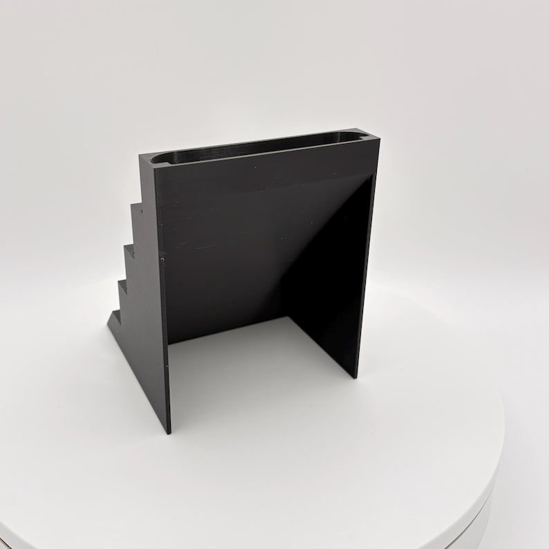 K&ouml;nnte beinhalten: Ein schwarzer, geometrischer Pr&auml;sentationsst&auml;nder mit einem Stufendesign auf einer Seite und einer flachen Oberfl&auml;che auf der anderen Seite. Der St&auml;nder hat eine rechteckige Oberseite und besteht aus einem soliden, undurchsichtigen Material. Das Design ist modern und minimalistisch.