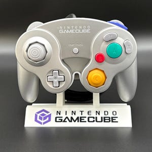 May include: Silver Nintendo GameCube controller with a white stand. The controller has a directional pad, analog stick, and colored buttons. The stand displays the Nintendo GameCube logo and text.