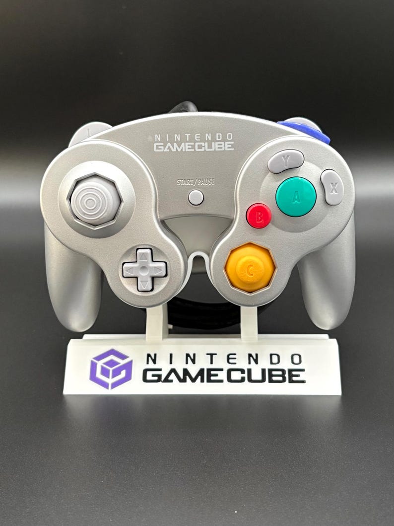 May include: Silver Nintendo GameCube controller with a white stand. The controller has a directional pad, analog stick, and colored buttons. The stand displays the Nintendo GameCube logo and text.