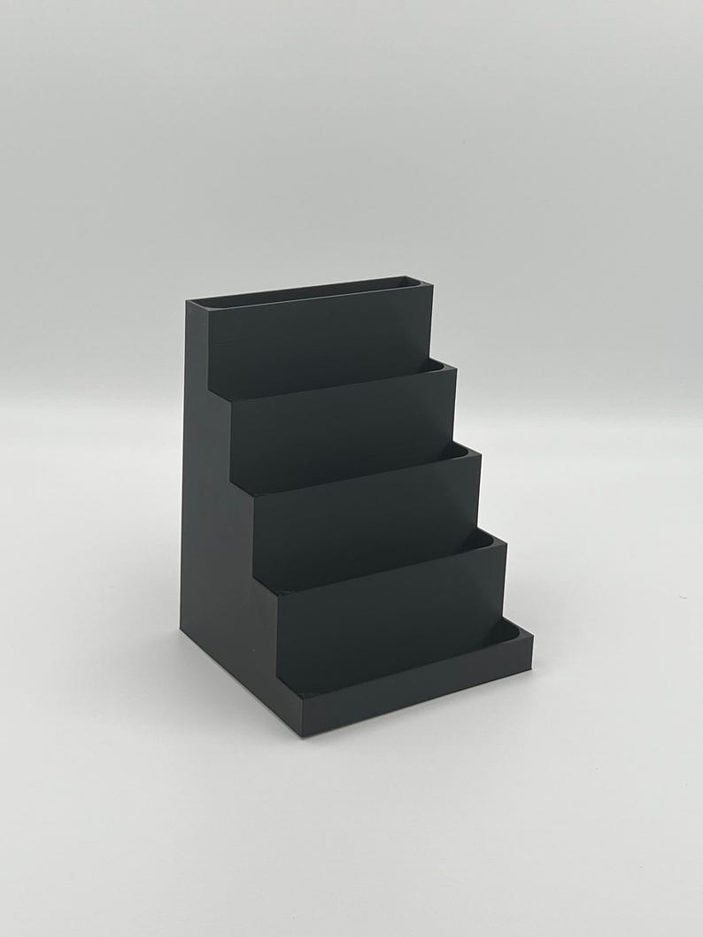 May include: A black plastic tiered desktop organizer with four compartments. The organizer is designed to hold business cards, pens, or other small office supplies.