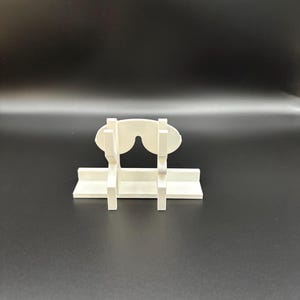 May include: A white, 3D-printed glasses holder. The holder is designed to resemble a pair of glasses, with a horizontal base and vertical supports. The glasses rest on the base, held in place by the supports. The holder is made of a solid, opaque white material.
