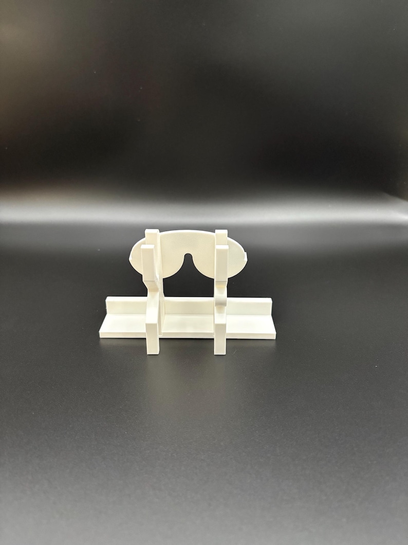 May include: A white, 3D-printed glasses holder. The holder is designed to resemble a pair of glasses, with a horizontal base and vertical supports. The glasses rest on the base, held in place by the supports. The holder is made of a solid, opaque white material.