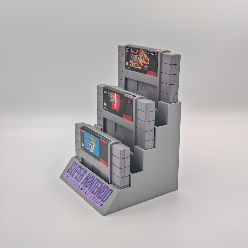 Puede incluir: Un soporte de pl&aacute;stico gris para exhibir cartuchos de juegos de Super Nintendo. El soporte tiene tres niveles y est&aacute; etiquetado como "SUPER NINTENDO ENTERTAINMENT SYSTEM".