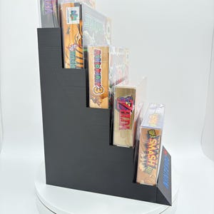 May include: A black, stepped display stand holding several video game boxes. The games include titles like "Zelda", "Star Fox 64", and "Super Smash Bros". The stand is made of a solid, matte black material and is designed to showcase the game boxes.