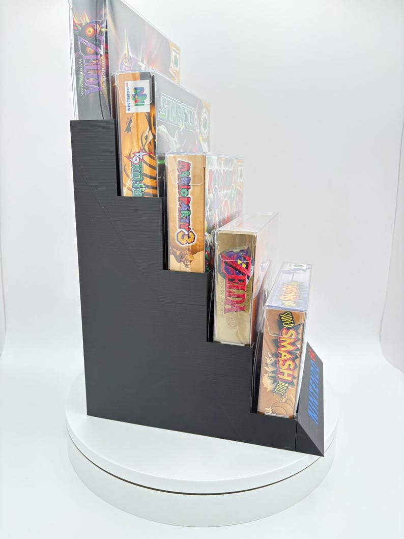 May include: A black, stepped display stand holding several video game boxes. The games include titles like "Zelda", "Star Fox 64", and "Super Smash Bros". The stand is made of a solid, matte black material and is designed to showcase the game boxes.
