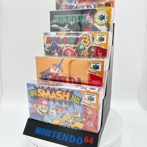 May include: A black display stand holding five Nintendo 64 video game boxes. The boxes feature game titles like "Super Smash Bros" and "Mario Party 3." The stand has the words "NINTENDO 64" in blue and red.