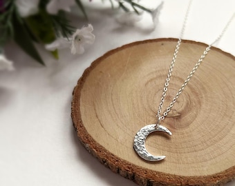 May include: A silver necklace with a hammered crescent moon pendant. The necklace is on a wooden surface.