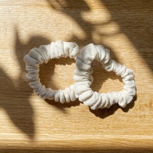 May include: Two off-white fabric hair scrunchies with a gathered, pleated texture. The circular hair accessories are made of a plain, light-coloured textile material and feature a soft, elasticised construction for securing hair in a ponytail or bun.