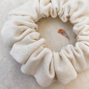 May include: A circular hair scrunchie made of off-white, textured fabric. The accessory features a gathered, pleated design with a soft, matte finish. The material has a visible weave, creating a simple, functional hair tie with a neutral colour.
