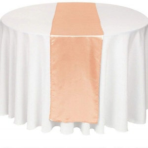 May include: A peach-colored satin table runner is centered on a round white tablecloth. The runner has a smooth, shiny texture and adds a touch of elegance. The table setting is ideal for weddings, parties, or special events.