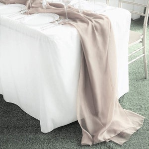 May include: A formal table setting with a white tablecloth and a flowing, taupe-colored table runner. White plates, silverware, and glassware are arranged on the table, suggesting a wedding or event.