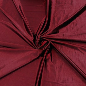 May include: A close-up shot of a deep burgundy fabric with a lustrous sheen. The fabric is gathered and folded, creating a swirling pattern of light and shadow. The material appears soft and drapes elegantly, suggesting it could be used for clothing or home decor.