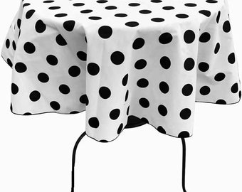 May include: A round white tablecloth with a pattern of large black polka dots. The tablecloth is draped over a table with black legs. The edges of the tablecloth are finished with a black trim.