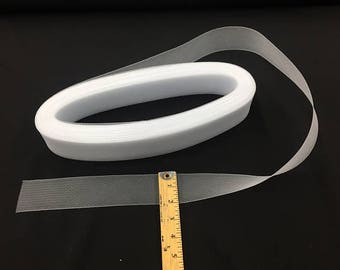 May include: A roll of white tulle ribbon with a fine mesh texture. The ribbon is partially unrolled, revealing its width. A wooden ruler is placed next to the ribbon for scale, showing measurements in inches.