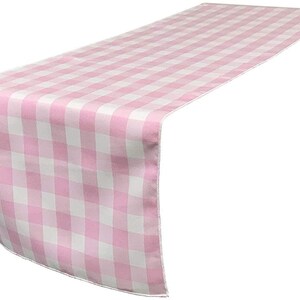 May include: A pink and white checkered table runner. The runner is made of a soft fabric and features a classic gingham pattern. The runner is designed to be placed on a table to add a decorative touch.