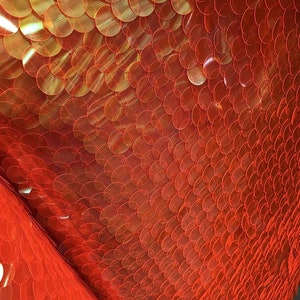 May include: Close-up of a vibrant red sequin fabric. The sequins are circular and overlap, creating a textured surface. The fabric is on a mesh backing, and the sequins reflect light, giving a shimmering effect. The fabric is folded, showing the texture and depth.