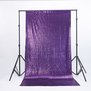 May include: A purple sequin backdrop hangs from a black metal stand. The backdrop is a solid color, with a shiny, reflective surface. The stand has three legs and is adjustable.