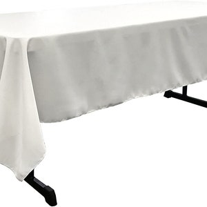 May include: A white rectangular tablecloth draped over a black folding table. The tablecloth is made of a smooth, opaque fabric, with the corners hanging down. The table legs are black.