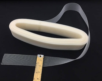 May include: A roll of off-white tulle ribbon, partially unrolled, with a section measured by a wooden ruler. The ribbon is on a black surface. The ruler shows measurements in inches.