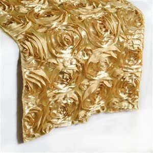 May include: Gold-colored table runner with a repeating rose pattern. The fabric has a textured appearance, with the roses creating a three-dimensional effect. The runner is draped over a white surface.