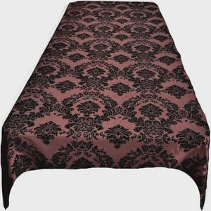 May include: A rectangular tablecloth featuring a repeating black damask pattern printed on a dusty rose pink fabric base. The fabric is draped to show a smooth surface with a consistent, symmetrical floral and scrollwork design throughout.