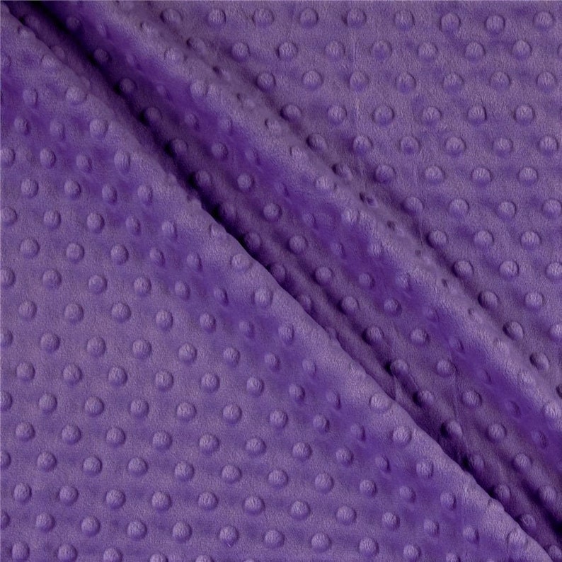 May include: Close-up of a soft, purple fabric with a textured, dimpled surface. The fabric is folded, creating shadows and highlighting the plush texture. The color is a vibrant, medium purple.
