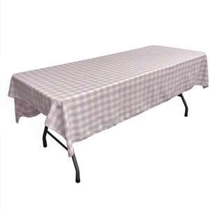 May include: A rectangular tablecloth in a pink and white gingham pattern covers a folding table. The tablecloth drapes over the edges of the table, which has black metal legs. The fabric has a classic, country-chic aesthetic.