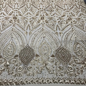 May include: A close-up of a cream-coloured fabric with an elaborate gold sequin pattern. The design features geometric shapes and stylised leaf-like motifs, creating a luxurious and ornate texture. The fabric is likely used for formal wear or decorative purposes.