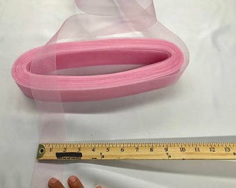 May include: A roll of pink tulle ribbon, partially unrolled, with a wooden ruler alongside. The ribbon is approximately 1 inch wide. The ruler shows measurements in inches. The tulle is a soft, sheer fabric, often used for crafts and decorations.