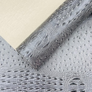 May include: A roll of gray faux leather with a crocodile skin texture is partially unrolled, revealing a sheet with an ostrich skin pattern. The material is a neutral gray color, suitable for various crafting projects.