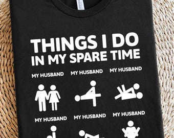 May include: Black t-shirt with white text that reads "THINGS I DO IN MY SPARE TIME." Below the text are stick figure illustrations and the words "MY HUSBAND" repeated. The shirt is made of a soft material.
