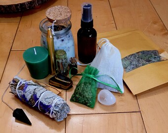 May include: A collection of green, white and brown items for a spiritual ritual. The items include a green candle, a white jar with a cork lid, a small bottle with a cork lid, a brown bottle with a spray top, a white pouch, a green pouch, a white and brown pouch, a white and purple bundle of sage, a black pendulum, a clear crystal, and a brown and gold stone.