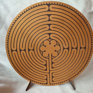 Op de afbeelding: Een rond houten labyrint met een bruine afwerking en donkerbruine geëtste paden. Het ontwerp toont concentrische cirkels en een centraal bloemmotief. Het labyrint wordt ondersteund door een kleine houten standaard.