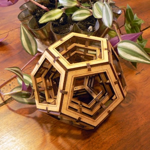 Pode incluir: Uma escultura geométrica de madeira castanha clara com um design complexo e em camadas. A escultura apresenta formas pentagonais interligadas. A imagem também inclui uma planta em vaso com folhas verdes e roxas.