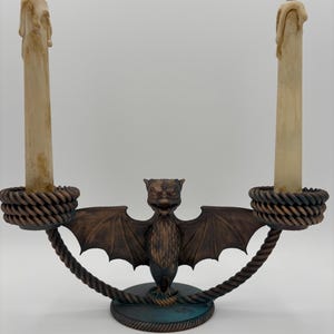 May include: A bronze-colored candelabra with a bat-like creature in the center. The candelabra holds two cream-colored candles with faux wax drips. The base and the arms of the candelabra have a rope-like design.