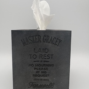 May include: A dark gray tissue box with the words "MASTER GRACEY LAID TO REST" and "Farewell" engraved on the front. White tissues are pulled from the top. The box also includes the text "NO MOURNING PLEASE AT HIS REQUEST."
