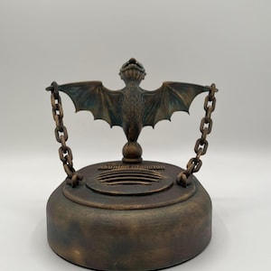 May include: Bronze-colored decorative bat-shaped object with outstretched wings, suspended by chains from a circular base. The base has the text "Haunted Mansion" on it. The bat has detailed wing and body features.